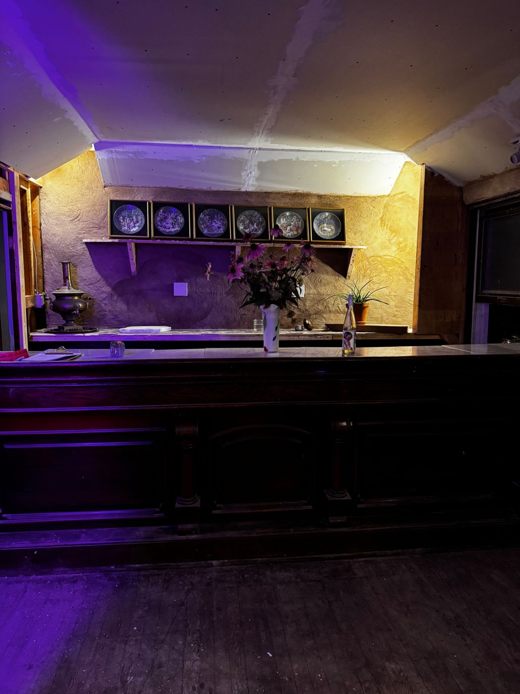 The Tavern's original wooden bar, lit in purple and gold, with framed plates on the plaster wall behind it